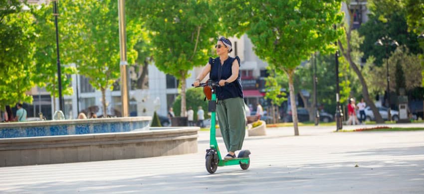 Mujer trasnportándose en un scooter eléctrico junto a las áreas verdes de una ciudad